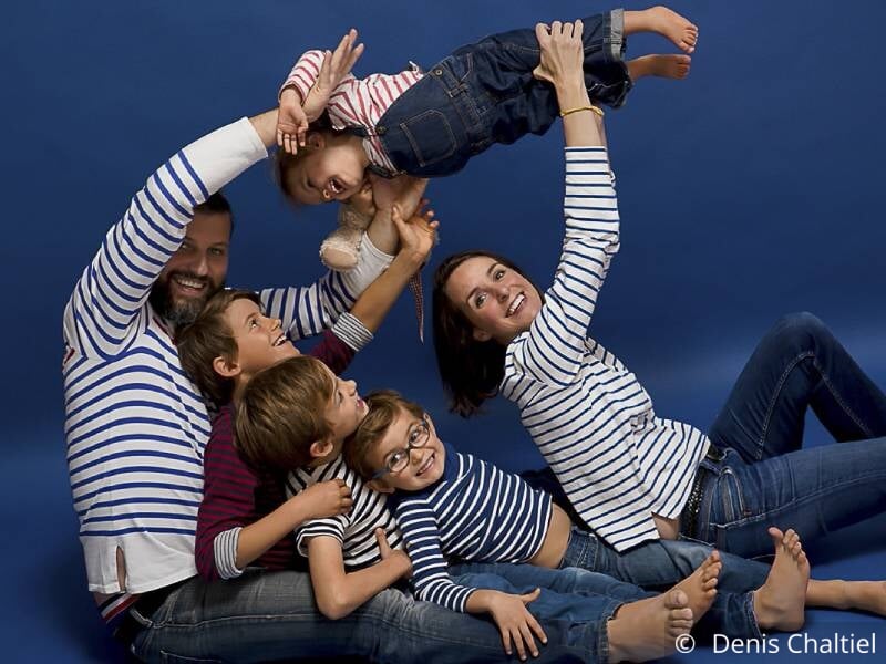 Photo de famille sur fond bleu, 2 parents et 4 enfants sourient, tous portent une marinière et un jean bleu.